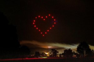 A heart shape is formed by red lights in the night sky above a park with trees and a lit building in the background, all part of a breathtaking drone show.