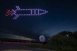 A mesmerizing drone show featured a formation of drones creating the image of a rocket in the night sky above a landscape with trees.
