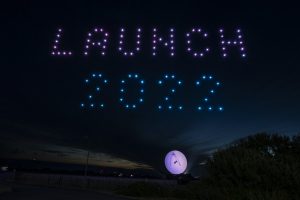 A night sky illuminated by a mesmerizing drone show forms the words "LAUNCH 2022" above a lit-up spherical object on the ground.