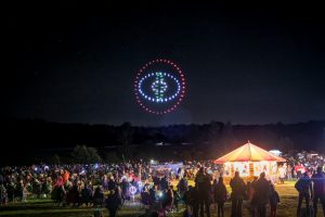 Nighttime festival with a crowd watching a carousel and an enchanting drone show, featuring a light display shaped like an eye in the sky.