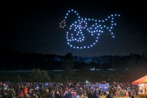 A crowd watches an impressive drone show forming an image of Snoopy against the night sky.