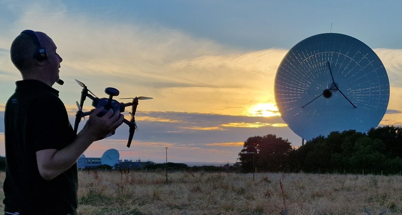 As the sun sets in a golden field, a person orchestrates an impressive drone show near large satellite dishes.