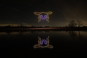 A dark night sky hosts a mesmerizing drone show, with lights forming a glowing Pikachu face, reflected on a calm lake surrounded by bare trees in the background.