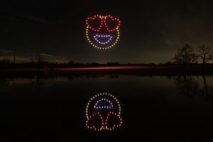 A night sky with a spectacular drone show featuring a smiley face with heart eyes, reflected in a lake surrounded by trees.