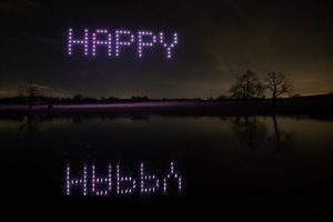 A drone light show in the night sky spells "HAPPY" with a reflection in a lake below, against a dark landscape background.