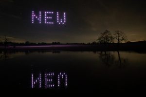 A night sky with an impressive drone light show spelling "NEW" and its reflection on a calm body of water, framed by silhouetted trees.
