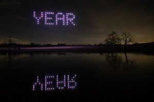 A spectacular drone light show unfolds as drones form the word "YEAR" in purple lights against a night sky, with trees and a lake reflecting the display below.