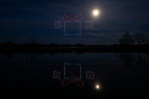 A drone light show illuminates the night sky and reflects on the water, forming a vibrant design under the moonlight.