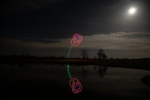 Colorful drone light show forms a rose shape with its reflection visible on a calm lake under a moonlit night sky.
