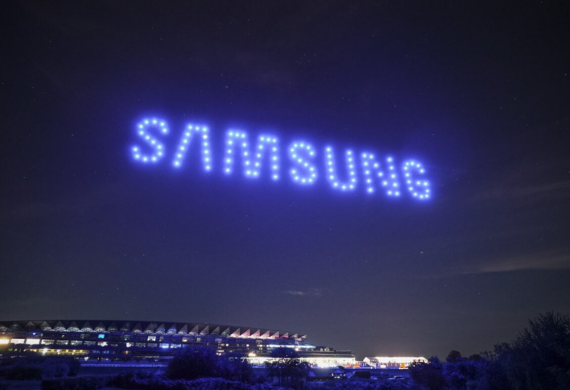 Blue lights from a droneswarm spell out "SAMSUNG" above a large building and trees, creating a stunning display in the night sky.