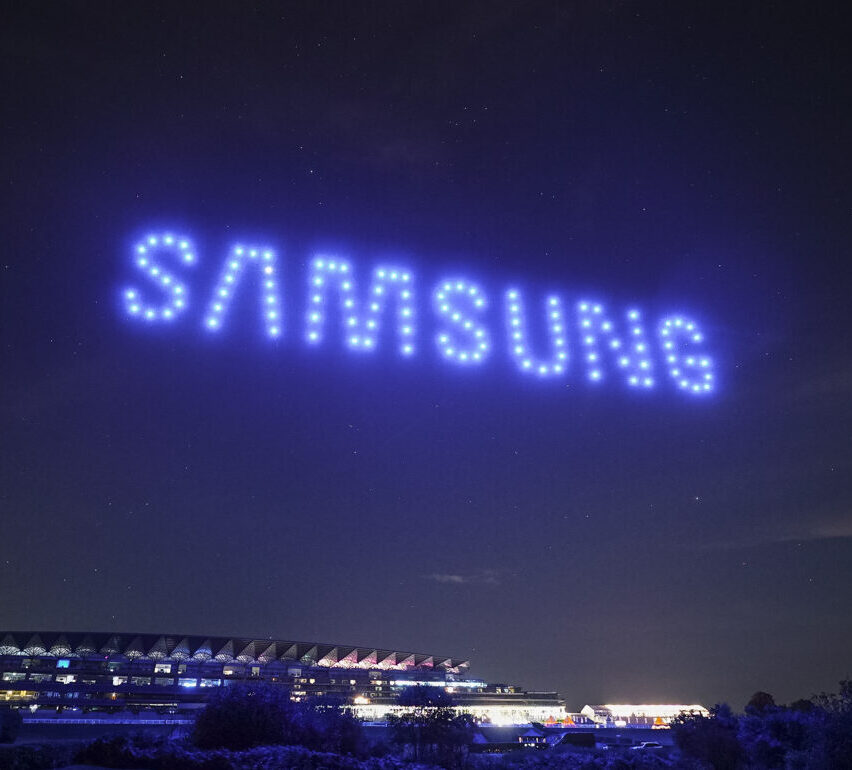 Blue drones from a Droneswarm create the word "SAMSUNG" in the night sky above a building with lights, showcasing the magic of drone light shows.