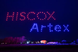 A dazzling drone light show lit up the night sky, spelling "HISCOX" in vivid red and "Artex" in stunning blue above a picturesque landscape.