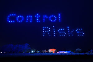 A nighttime sky display features a mesmerizing drone light show, with blue drones forming the words "Control Risks" above a landscape adorned with trees and lights.