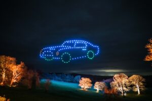 In a mesmerizing drone light show, drones form a glowing blue and green car in the night sky above a field with illuminated trees.