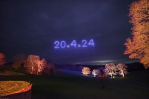 A nighttime landscape with illuminated trees and a sky displaying "20.4.24" in blue lights formed by a mesmerizing drone light show.