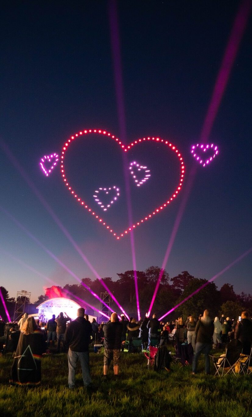 A crowd watches in awe as the Floyd Drone Show lights up the night sky, forming glowing pink hearts above an outdoor event.