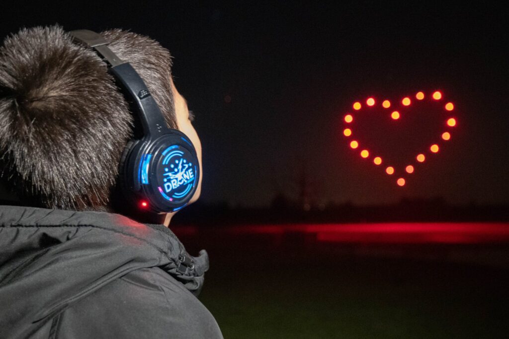 A person wearing headphones looks at a red heart shape made of lights in the night sky.
