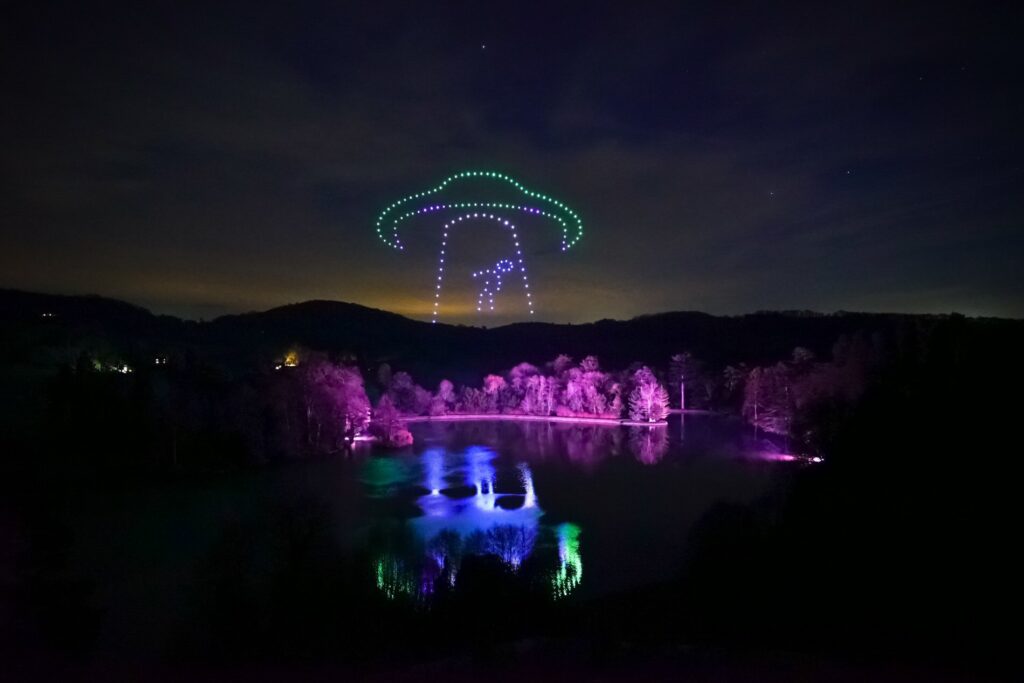 A nighttime sky display: lights form a colorful UFO shape over a forest and lake, reflecting on the water.