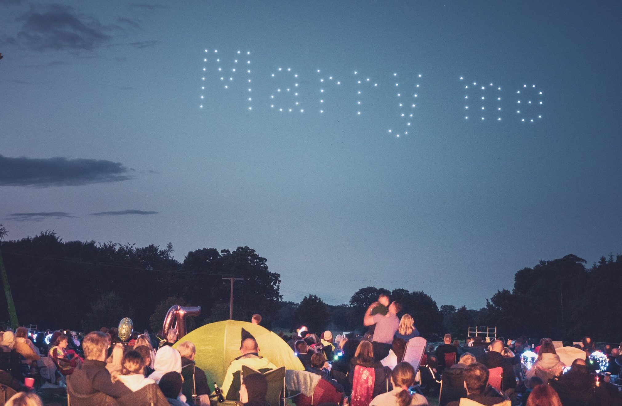 A crowd watches as drones form the words "Marry me" in lights in the evening sky.