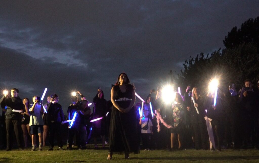 A woman stands outside at night as a crowd behind her holds glowing lights and flashes from phones. Drone Gender Reveal