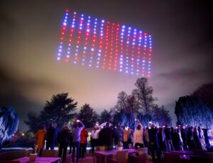 A crowd watches a nighttime drone light show with red and blue lights forming shapes in the sky.