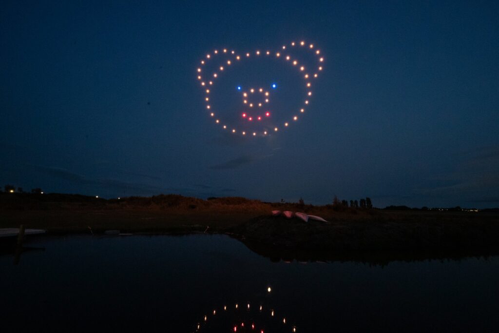 Drones form a glowing bear face in the night sky, reflected in the calm water below.