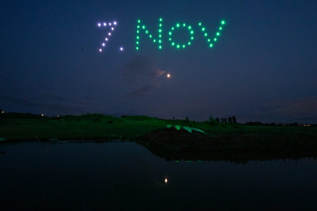 Drones form "7 NOV" in lights over a grassy landscape at dusk, reflected in a calm body of water.