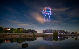 A formation of drones creates a glowing Reddit mascot in the night sky above a lake and trees.