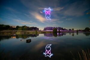 A colorful owl made of lights in the sky, reflected on a calm lake at night with trees in the background.