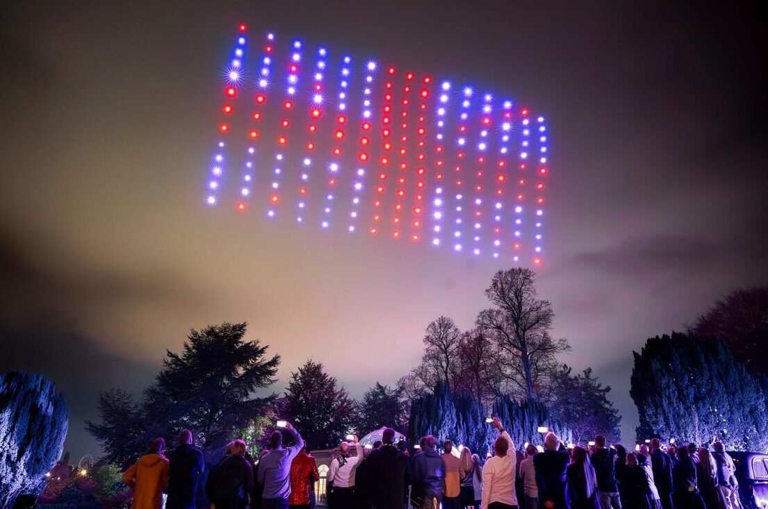 A crowd watches as a DroneSwarm forms red and blue patterns, resembling the DroneSwarm Flag, in the night sky above trees.