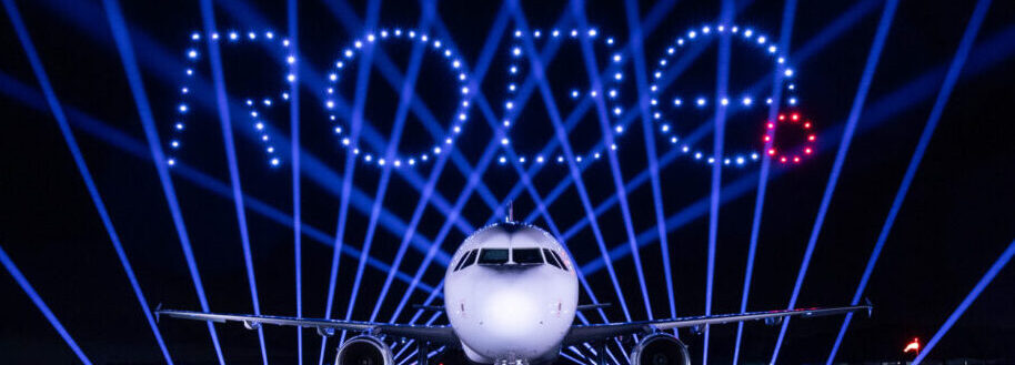 A plane with “Rome” spelled in lights above it, possibly crafted by a DroneSwarm, and blue beams radiating in the background at night.