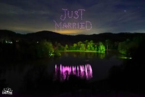 A nighttime lake scene with drones in the sky spelling out "JUST MARRIED" above illuminated trees.