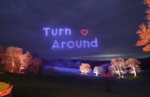 Trees lit at night; drones form "Turn Around" and a heart shape in blue and red lights above.