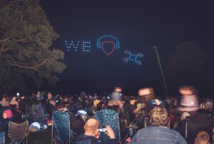 A crowd watches drones form "We ♥ X" with lights in the night sky at an outdoor event.