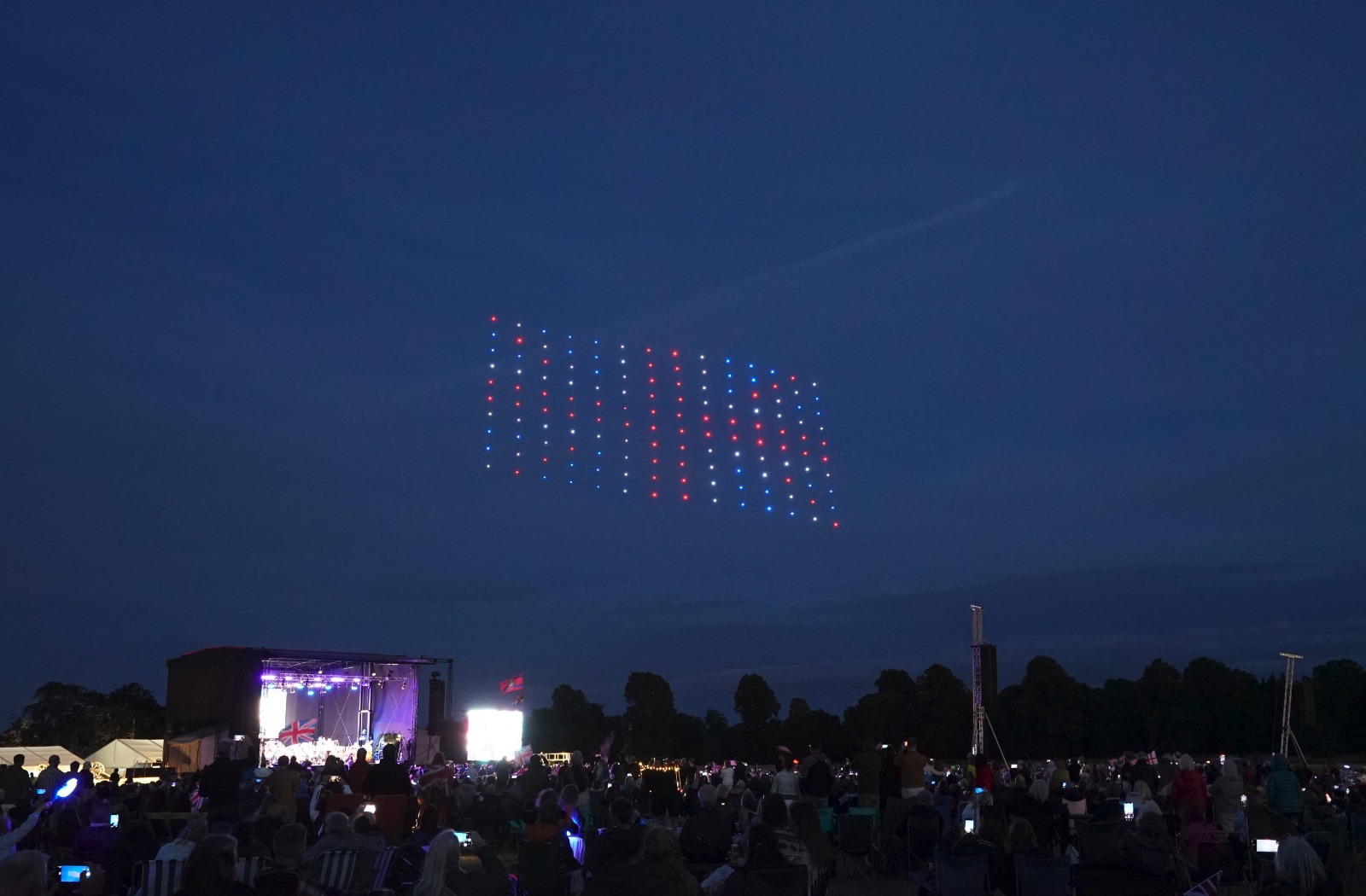 A crowd watches at Blenheim Palace as drones form a flag pattern with red, white, and blue lights in the night sky.