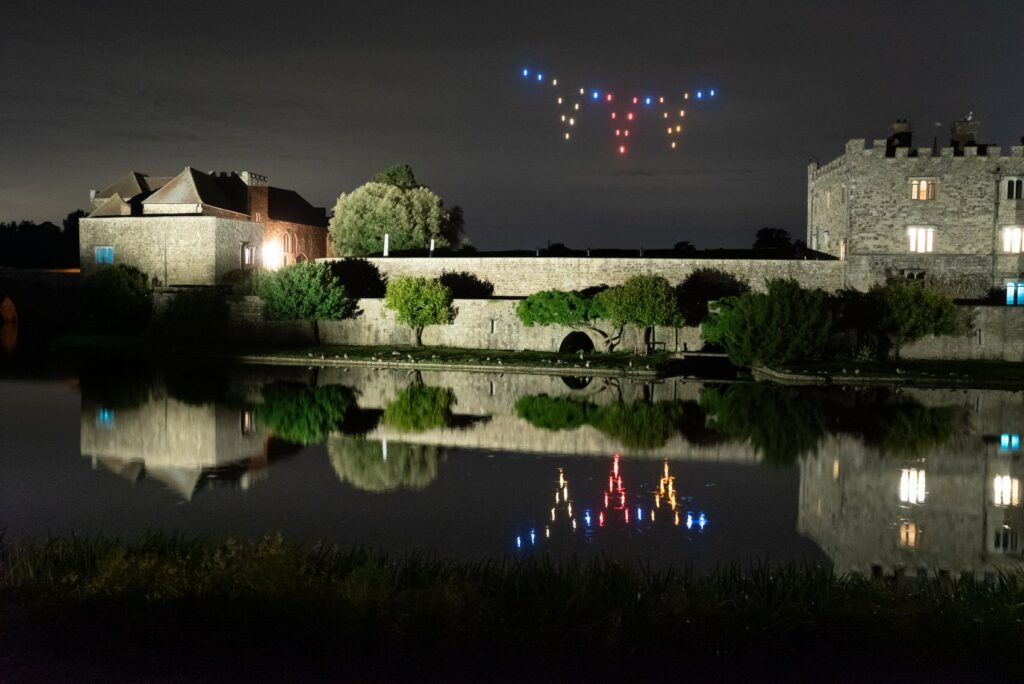 A nighttime castle scene with colored lights in the sky and their reflections in the water below.