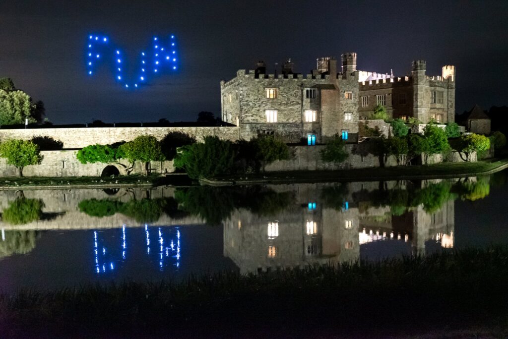 A castle at night with blue drone lights forming "FWA" in the sky, reflected in a calm moat.
