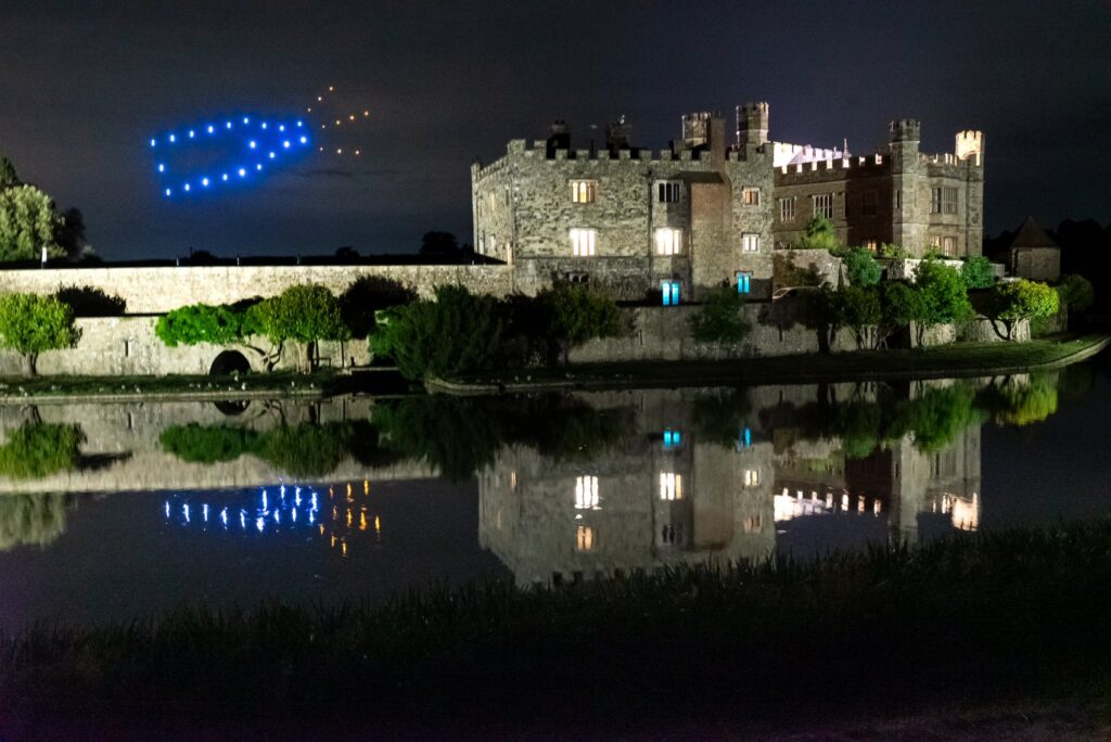 A castle at night with blue and yellow drones forming a pattern in the sky, reflected in a calm moat.