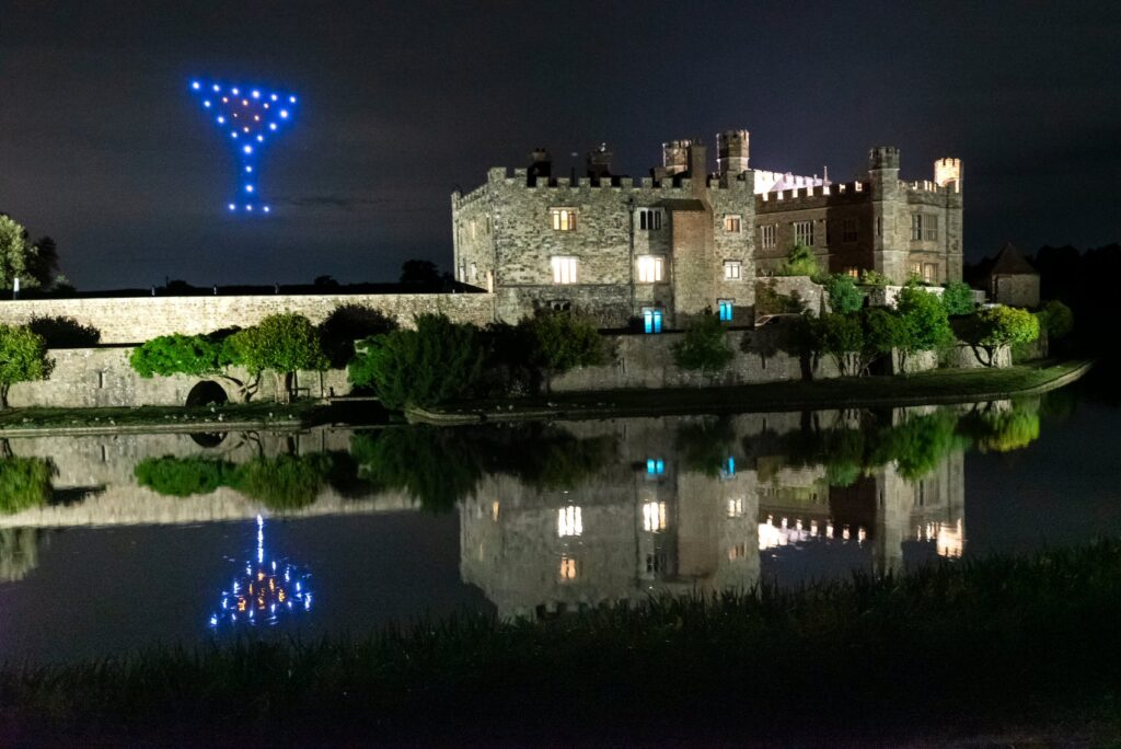 A castle at night with a martini glass shape formed by blue drones in the sky, reflected in the water.