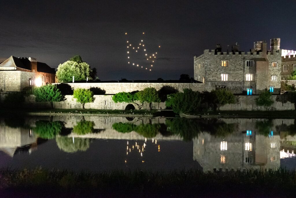 A nighttime castle scene with drones forming a heart in the sky, reflected in the water below.