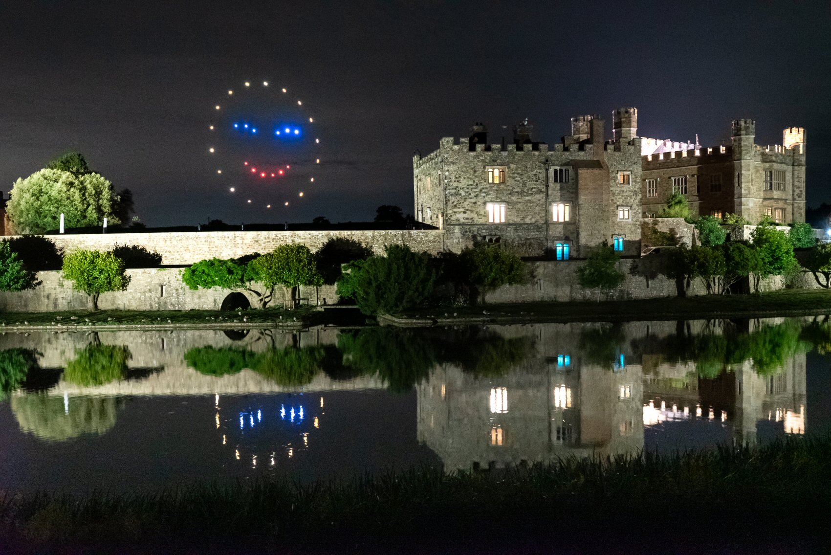 A smiley face made of drones glows above a lit castle at night, reflecting in the water below.