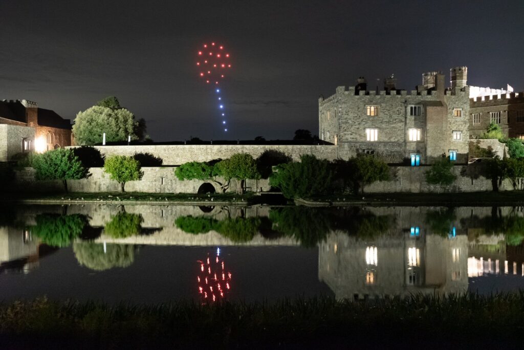 Drones form a red smiley face in the night sky above a castle, reflected in a calm moat.
