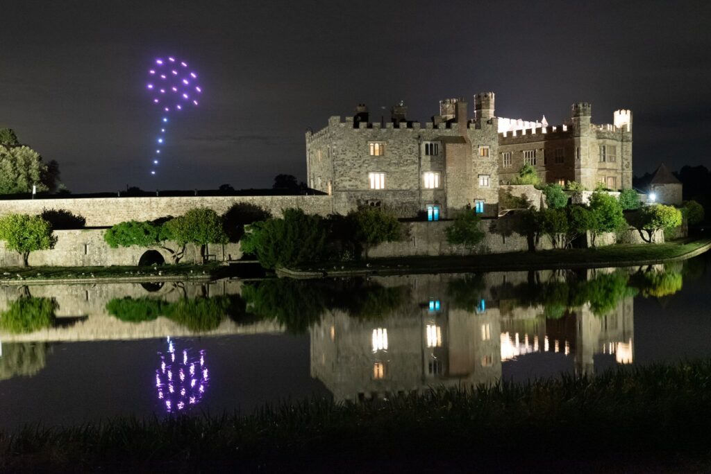 A stone castle at night with purple fireworks in the sky reflected in a calm moat.