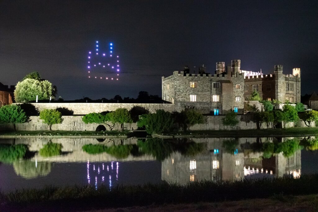 A castle by a river at night with a cat face made of drones glowing in the sky.