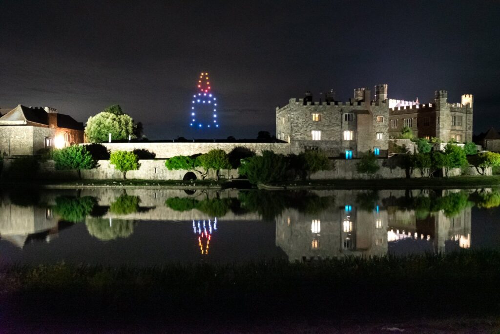 A castle at night is reflected in water with a rocket shape made of colorful lights in the sky.