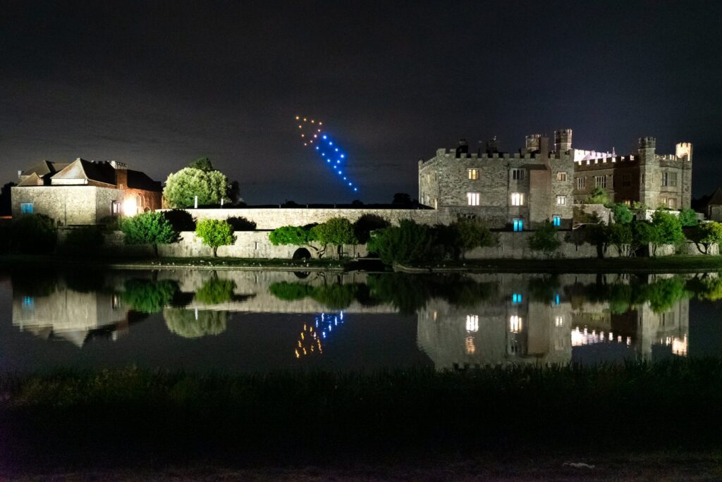 A castle at night is reflected in water, with blue and yellow lights forming shapes in the sky above.