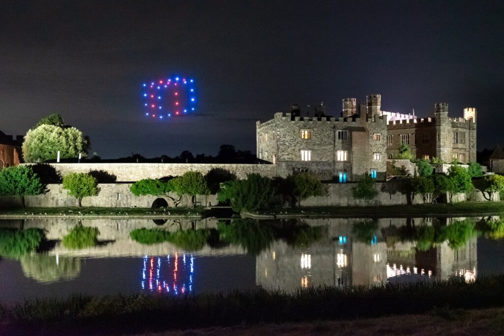 A castle at night with drones forming a "10" in blue and red lights above it, reflected in the water.