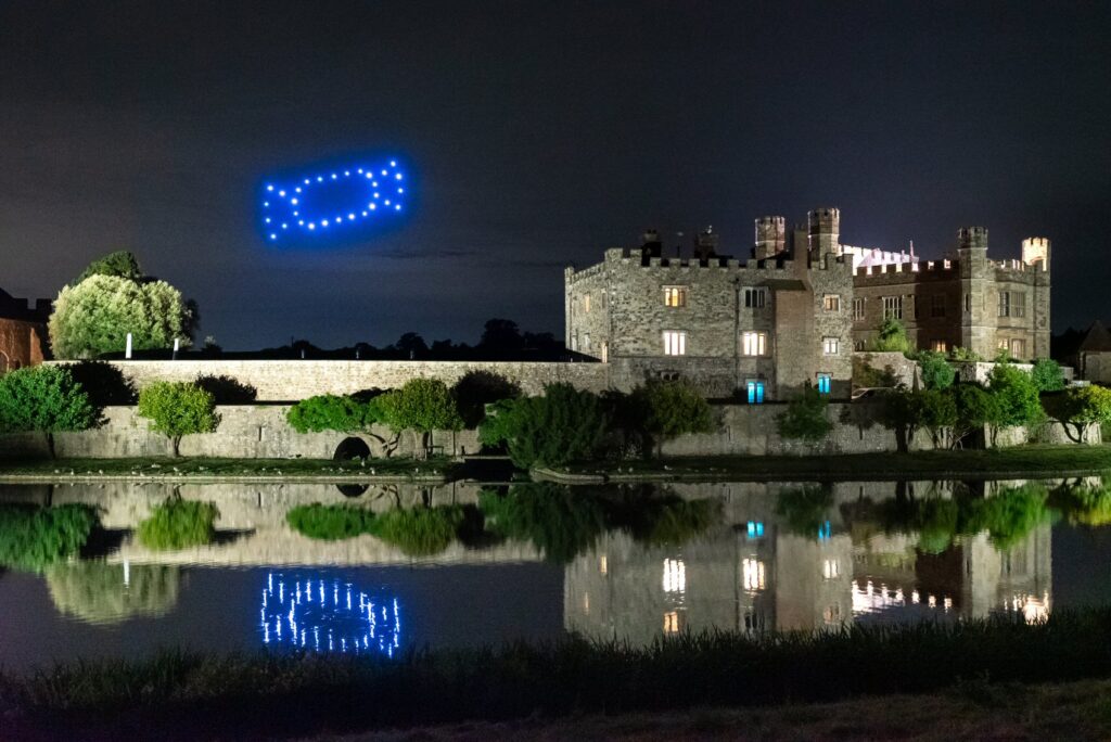 A lit-up castle at night with drones forming shapes in the sky, reflected in a calm river.