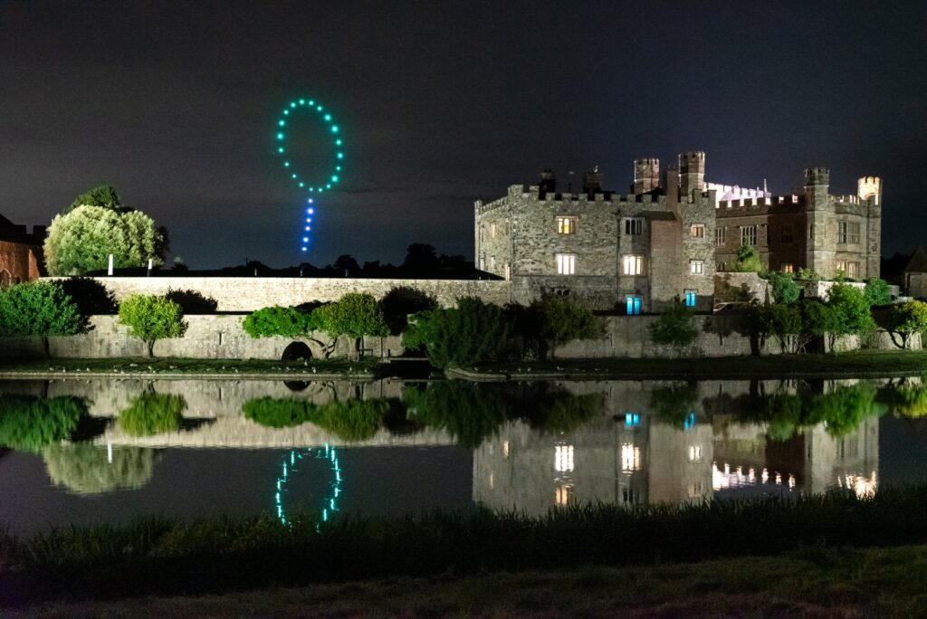 Castle at night with green and blue drone lights forming a question mark in the sky, reflected in the water.
