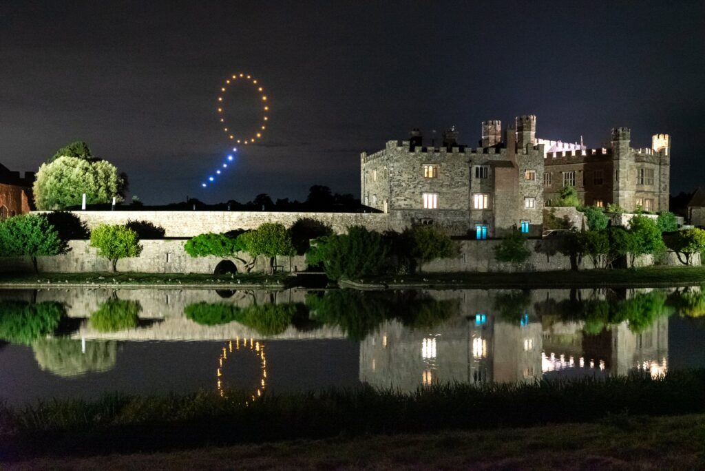 A lit castle at night is reflected in water with a circle of drones glowing in the sky above.